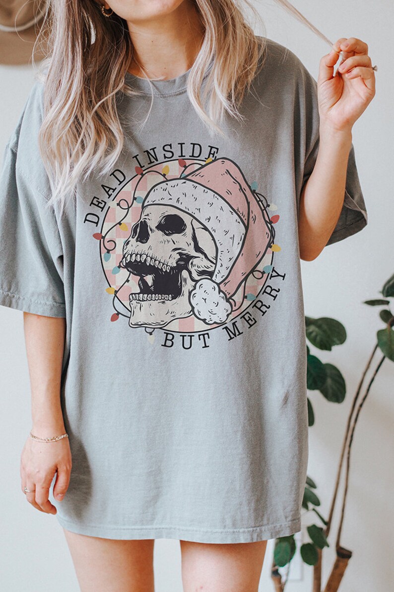 May include: A light blue t-shirt with a graphic of a skull wearing a Santa hat and surrounded by Christmas lights. The text "Dead Inside But Merry" is printed on the shirt.