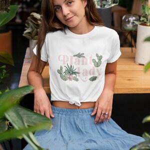 May include: A white t-shirt featuring the text "Plant Lady" in pink lettering and a graphic of various cacti and succulents. The model is wearing a blue skirt and is surrounded by plants.