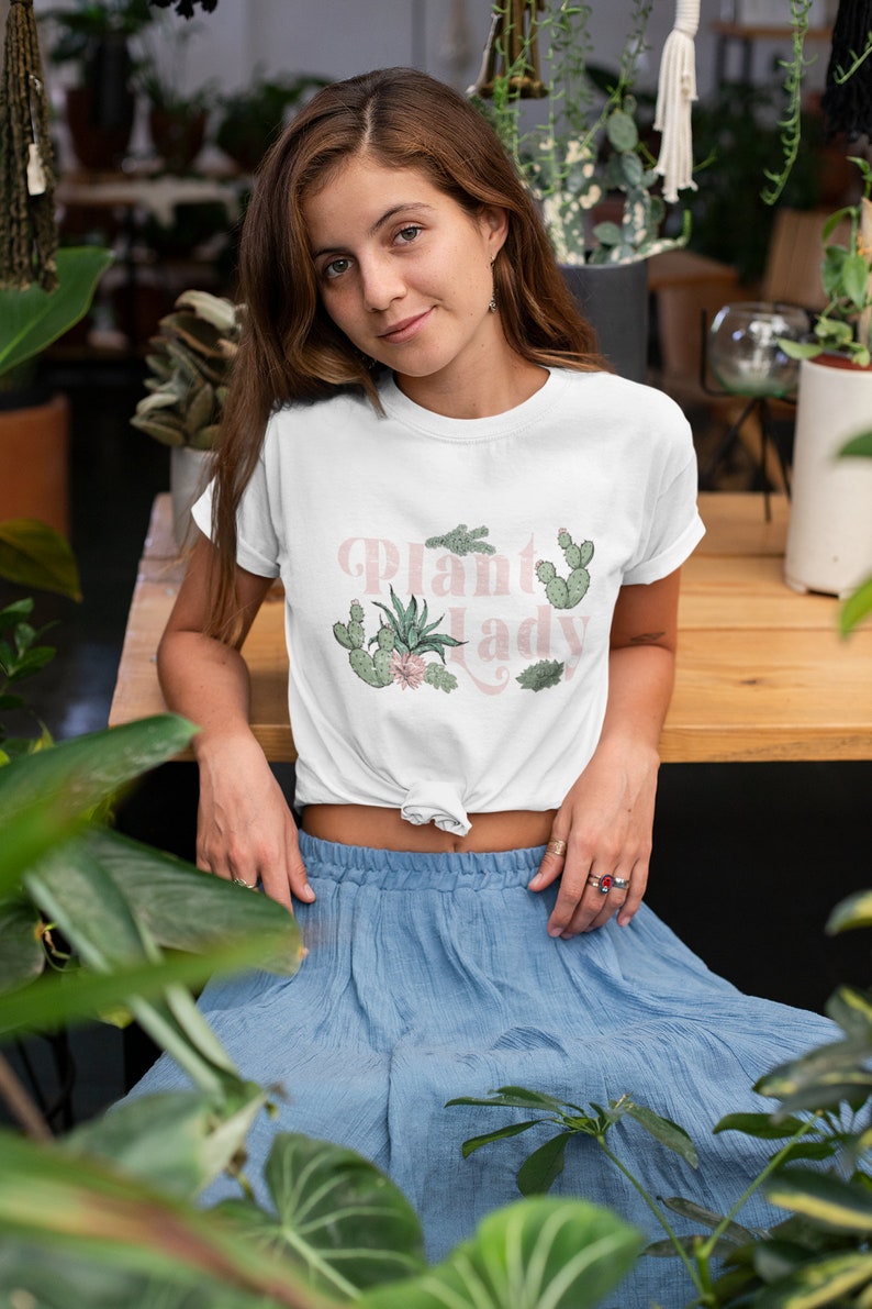 May include: A white t-shirt featuring the text "Plant Lady" in pink lettering and a graphic of various cacti and succulents. The model is wearing a blue skirt and is surrounded by plants.