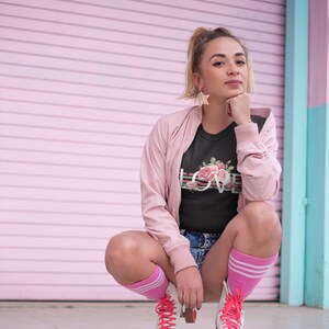 May include: A woman wearing a black t-shirt with a floral design and the word "LOVE" printed on it, pink socks, and white roller skates with pink laces. She is sitting on the ground in front of a pink and blue wall.