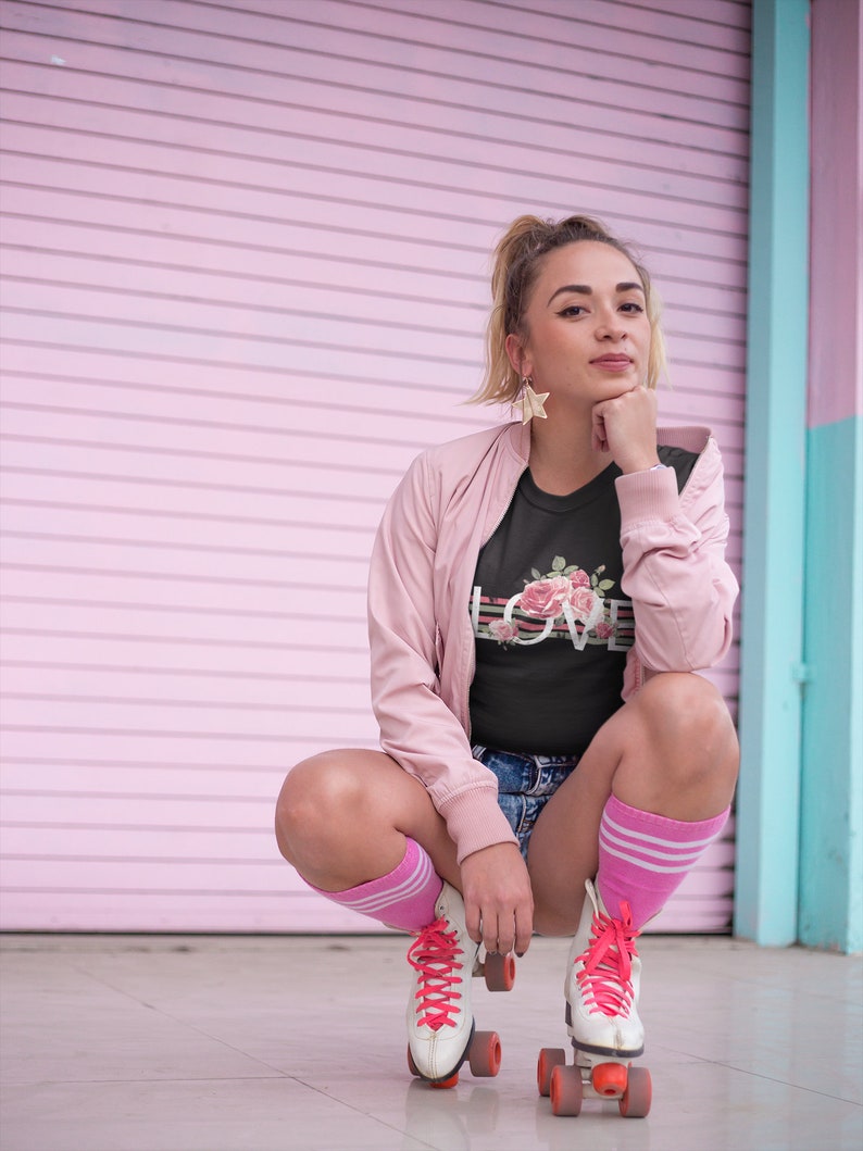May include: A woman wearing a black t-shirt with a floral design and the word "LOVE" printed on it, pink socks, and white roller skates with pink laces. She is sitting on the ground in front of a pink and blue wall.
