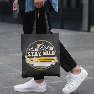 May include: A black canvas tote bag with a yellow and white graphic of mountains and daisies. The graphic says "Stay Wild".
