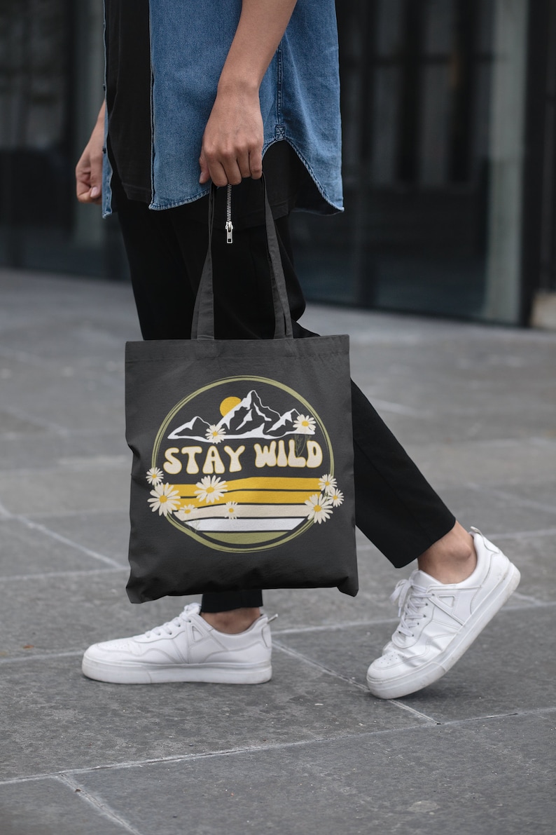 May include: A black canvas tote bag with a yellow and white graphic of mountains and daisies. The graphic says "Stay Wild".
