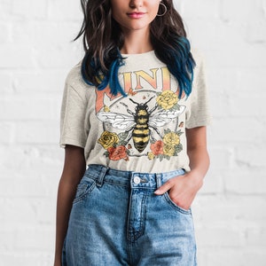May include: A light gray t-shirt with a vintage-style graphic of a bee surrounded by flowers. The graphic includes the word "KIND" in a yellow and orange gradient.
