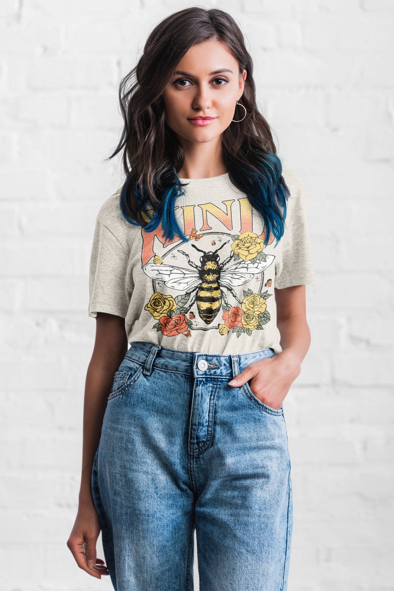 May include: A light gray t-shirt with a vintage-style graphic of a bee surrounded by flowers. The graphic includes the word "KIND" in a yellow and orange gradient.