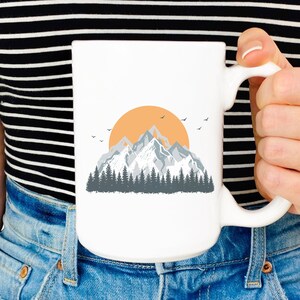 May include: White ceramic mug with a graphic of a mountain range with a sunset behind it. The mountains are gray and the sunset is orange.
