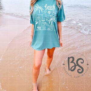 Może przedstawiać: Turkusowy t-shirt z białym nadrukiem przedstawiającym palmę, parasol plażowy, deskę surfingową i napis "Beach Vibes". Nadruk zawiera r&oacute;wnież tekst "Sunshine, Catch Wave, Relax, Eat Beach Sleep Repeat".