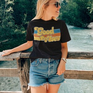 May include: A woman wearing a black t-shirt with the text "Good Vibes" in a retro font and blue denim shorts. She is standing on a wooden bridge over a river.