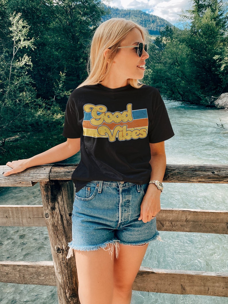 May include: A woman wearing a black t-shirt with the text "Good Vibes" in a retro font and blue denim shorts. She is standing on a wooden bridge over a river.