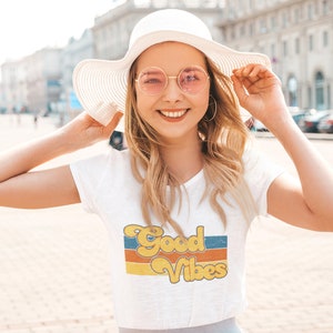 May include: A woman wearing a white t-shirt with a retro-style graphic that says "Good Vibes" in yellow and orange letters. She is also wearing a white sun hat and pink round sunglasses.