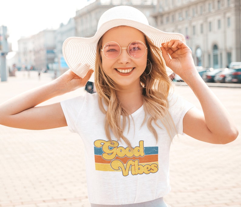 May include: A woman wearing a white t-shirt with a retro-style graphic that says "Good Vibes" in yellow and orange letters. She is also wearing a white sun hat and pink round sunglasses.