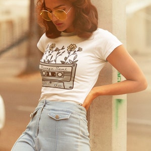 May include: A woman wearing a white t-shirt with a vintage cassette tape design and yellow flowers. The shirt says "Vintage Soul". She is wearing light blue jeans and has her hair styled in a retro look.