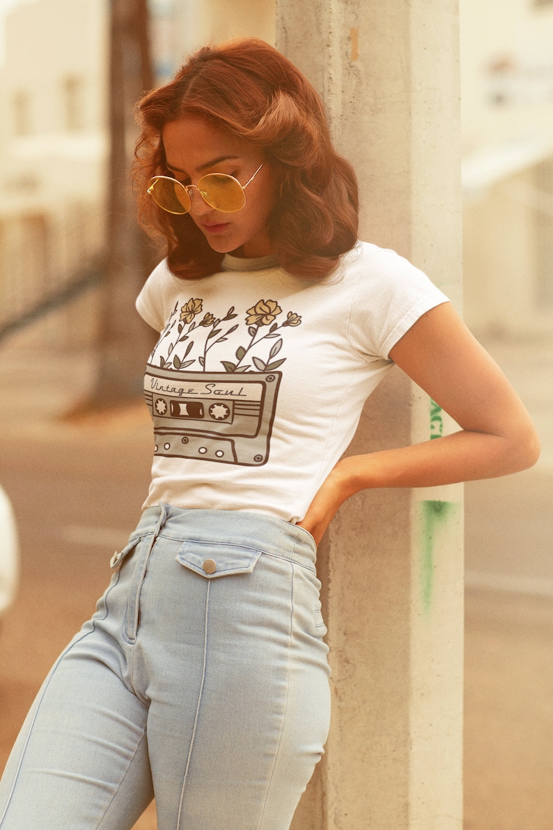 May include: A woman wearing a white t-shirt with a vintage cassette tape design and yellow flowers. The shirt says "Vintage Soul". She is wearing light blue jeans and has her hair styled in a retro look.