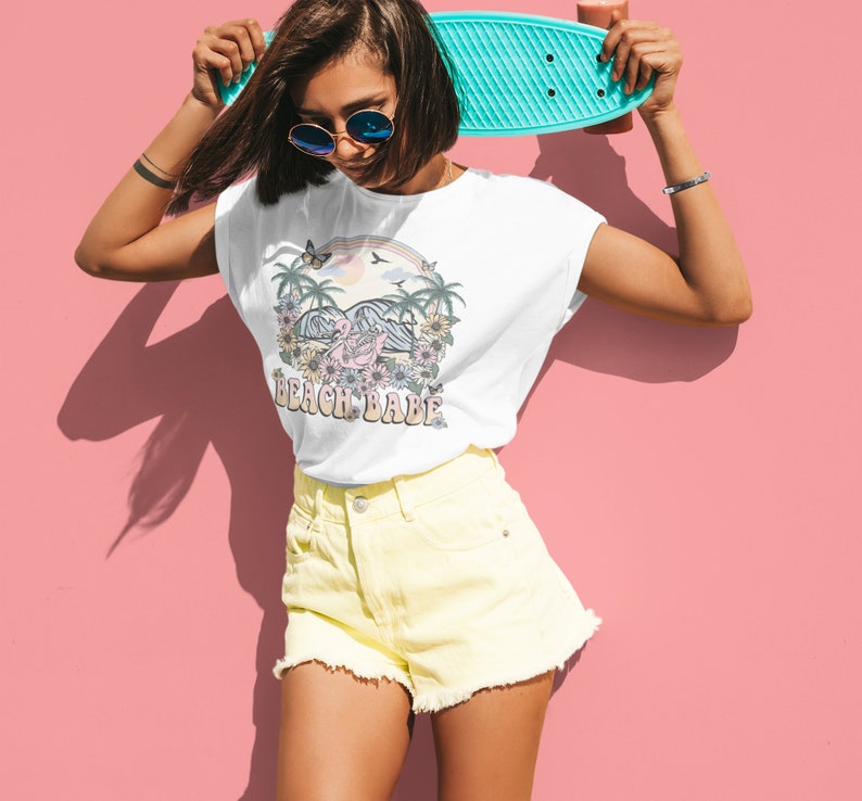 Op de afbeelding: Wit T-shirt met een afbeelding van een roze flamingo op een strand met palmbomen en de tekst "Beach Babe".