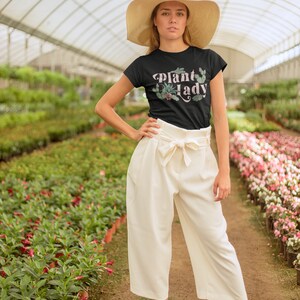 May include: A woman wearing a black t-shirt with the text "Plant Lady" in pink lettering and a green plant graphic. She is wearing a wide-brimmed straw hat and white trousers. She is standing in a greenhouse with rows of plants.