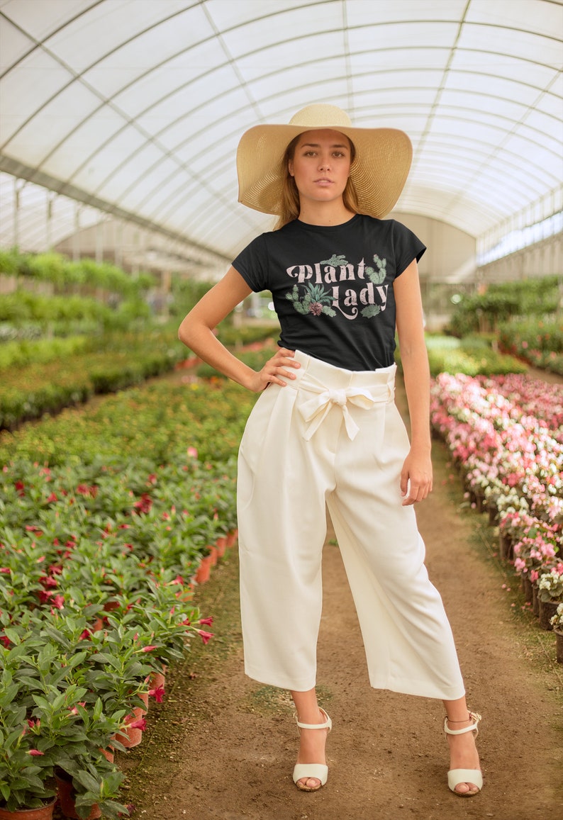 May include: A woman wearing a black t-shirt with the text "Plant Lady" in pink lettering and a green plant graphic. She is wearing a wide-brimmed straw hat and white trousers. She is standing in a greenhouse with rows of plants.
