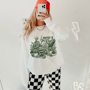 May include: White long-sleeve sweatshirt with a green skeleton graphic holding a cup of coffee and surrounded by plants. The text "Coffee & Plants" is printed on the sweatshirt.