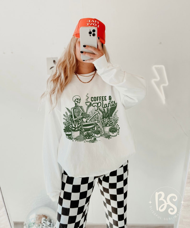 May include: White long-sleeve sweatshirt with a green skeleton graphic holding a cup of coffee and surrounded by plants. The text "Coffee & Plants" is printed on the sweatshirt.