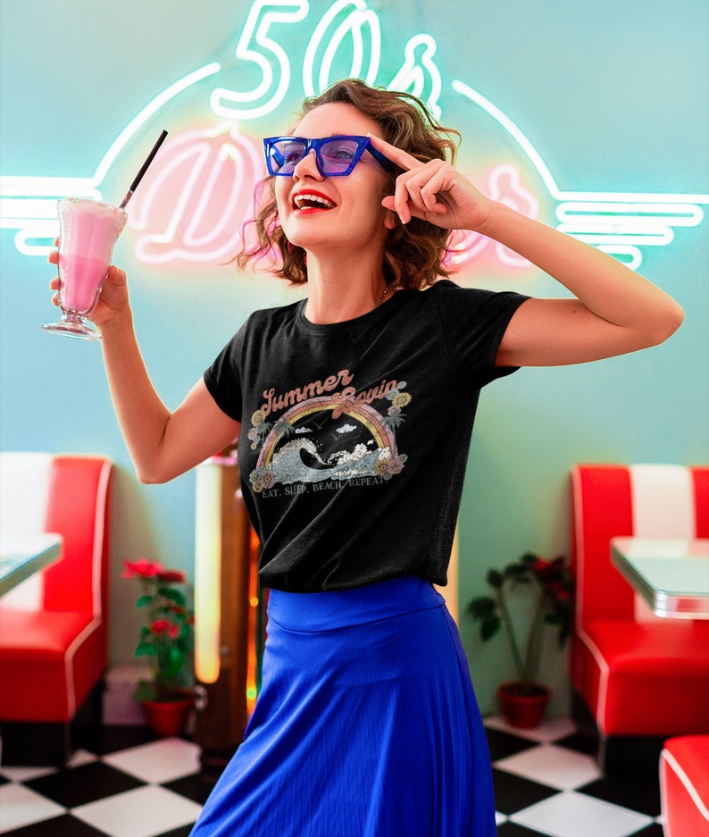May include: A woman wearing a black t-shirt with the words "Summer Loving" above a rainbow and wave graphic. She is wearing blue glasses and a blue skirt, holding a pink milkshake. The background features a neon sign.