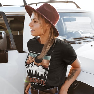 May include: A woman wearing a black t-shirt with a vintage-style graphic of a mountain range and the text "Yosemite National Park". She is wearing a brown hat and blue jeans.