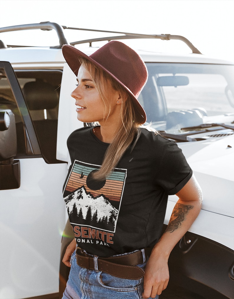 May include: A woman wearing a black t-shirt with a vintage-style graphic of a mountain range and the text "Yosemite National Park". She is wearing a brown hat and blue jeans.