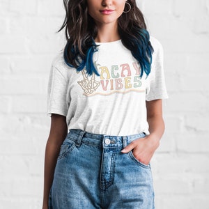 May include: A white t-shirt with a colorful graphic that says "Vacay Vibes" and features a skeleton hand making a peace sign.