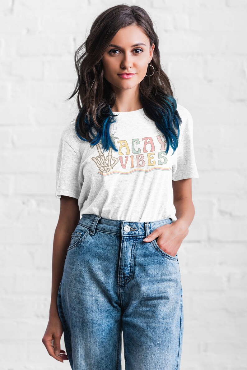 May include: A white t-shirt with a colorful graphic that says "Vacay Vibes" and features a skeleton hand making a peace sign.