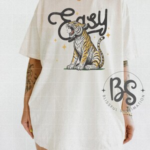 May include: White t-shirt with a graphic of a roaring tiger and the word "Easy" in a vintage style font. The shirt also has a small logo that says "Blissful Sublimation".