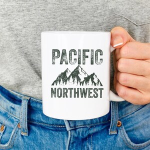 May include: White ceramic mug with a black and green graphic of mountains and trees. The text "PACIFIC NORTHWEST" is printed in black on the mug.