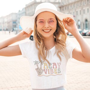 May include: White t-shirt with a colorful graphic of a skeleton hand giving the peace sign with palm trees and the text "Vacay Vibes".