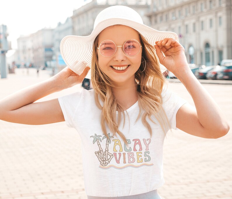 May include: White t-shirt with a colorful graphic of a skeleton hand giving the peace sign with palm trees and the text "Vacay Vibes".