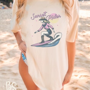 Casual t-shirt with a Surfing Cowgirl print, featuring a cowgirl surfing a wave with the text "Sunset Rider," ideal for beachwear and casual outfits