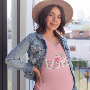 May include: A pink maternity top featuring a floral design and the word "LOVE" in white lettering. The top has a V-neck and is worn by a person in a denim jacket and a brown hat.