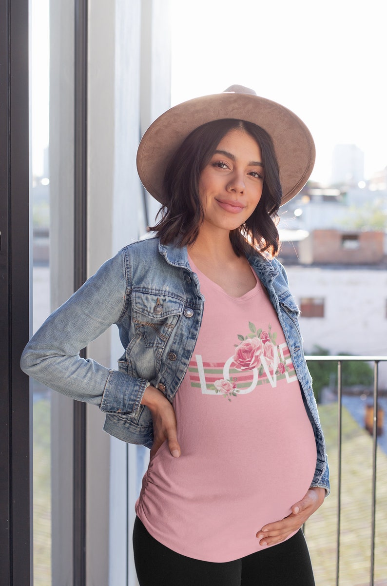 May include: A pink maternity top featuring a floral design and the word "LOVE" in white lettering. The top has a V-neck and is worn by a person in a denim jacket and a brown hat.