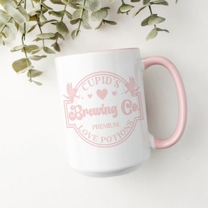 May include: White ceramic mug with a pink rim and handle. The mug features a pink graphic of a logo that says "Cupid's Brewing Co. Premium Love Potions".