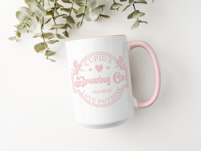 May include: White ceramic mug with a pink rim and handle. The mug features a pink graphic of a logo that says "Cupid's Brewing Co. Premium Love Potions".