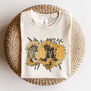 May include: A white t-shirt with a graphic of the word "MAMA" in a retro font surrounded by sunflowers and greenery.