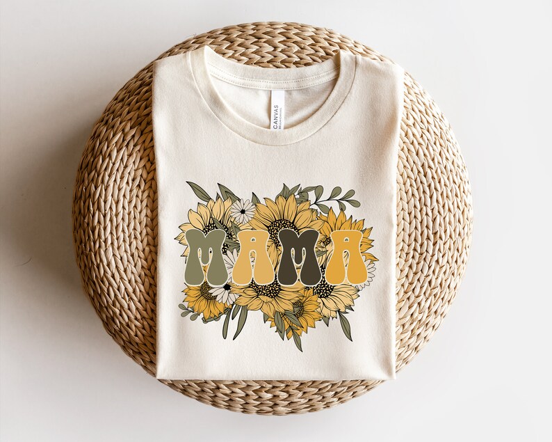 May include: A white t-shirt with a graphic of the word "MAMA" in a retro font surrounded by sunflowers and greenery.