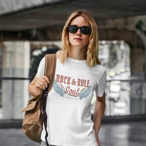 Puede incluir: Una camiseta blanca con un estampado gr&aacute;fico de alas y el texto "Rock & Roll Soul" con las palabras "make some noise" debajo.