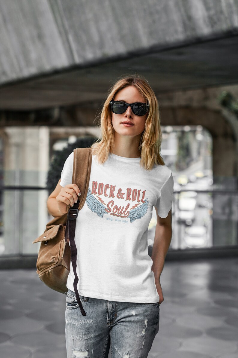Puede incluir: Una camiseta blanca con un estampado gr&aacute;fico de alas y el texto "Rock & Roll Soul" con las palabras "make some noise" debajo.