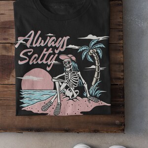 May include: Black t-shirt with a graphic of a skeleton wearing sunglasses and a hat sitting on a beach with a palm tree and the text "Always Salty".