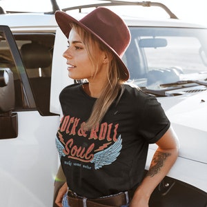 Puede incluir: Una mujer con una camiseta negra con un gr&aacute;fico de estilo vintage que dice "Rock & Roll Soul" con alas y el texto "make some noise". Tambi&eacute;n lleva un sombrero fedora marr&oacute;n, pantalones vaqueros azules y un cintur&oacute;n marr&oacute;n.