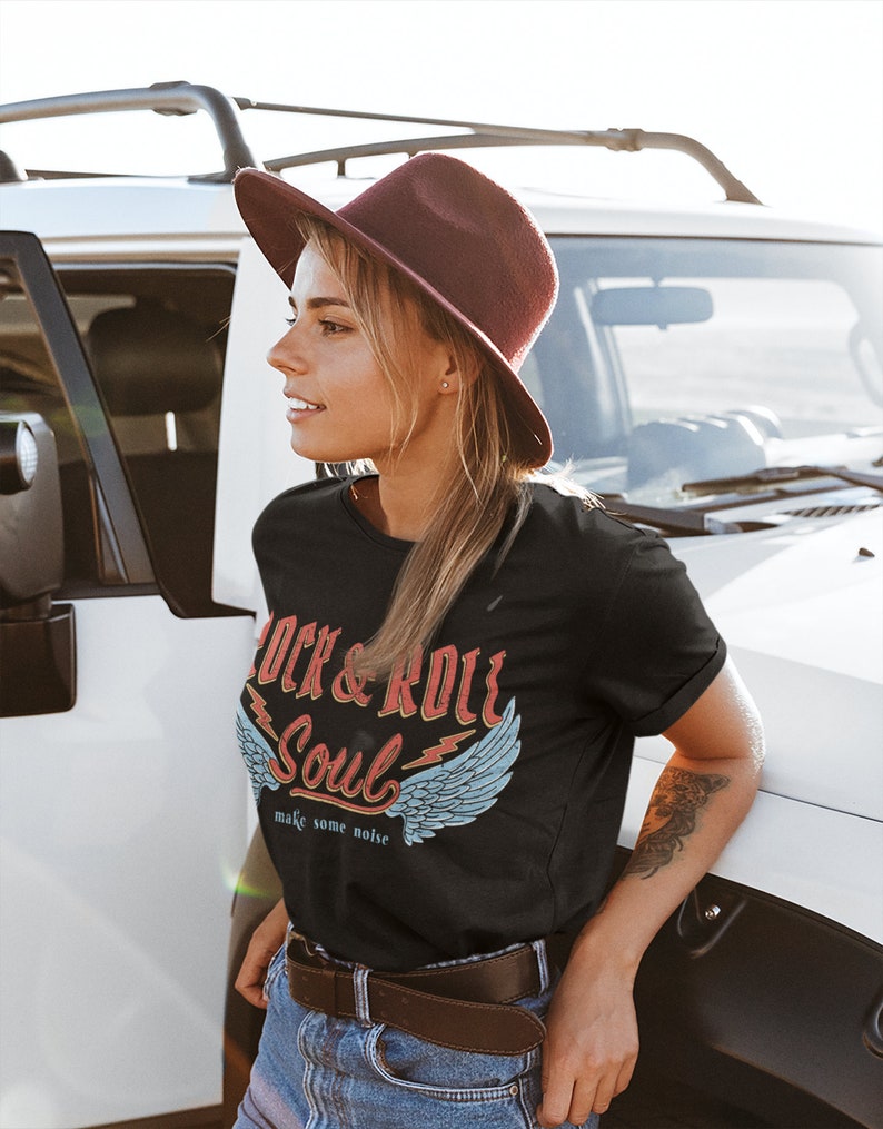 Puede incluir: Una mujer con una camiseta negra con un gr&aacute;fico de estilo vintage que dice "Rock & Roll Soul" con alas y el texto "make some noise". Tambi&eacute;n lleva un sombrero fedora marr&oacute;n, pantalones vaqueros azules y un cintur&oacute;n marr&oacute;n.