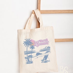 May include: A white canvas tote bag with a blue and pink graphic design of a beach scene with palm trees, a sunset, and a beach chair. The text "Saltwater and Sunshine" is printed on the bag.