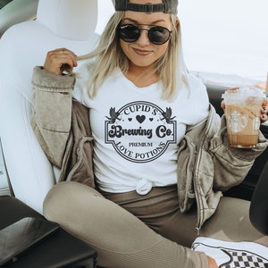 May include: White t-shirt with a black graphic that says "Cupid's Brewing Co. Premium Love Potions". The graphic features a heart and two winged cupids.