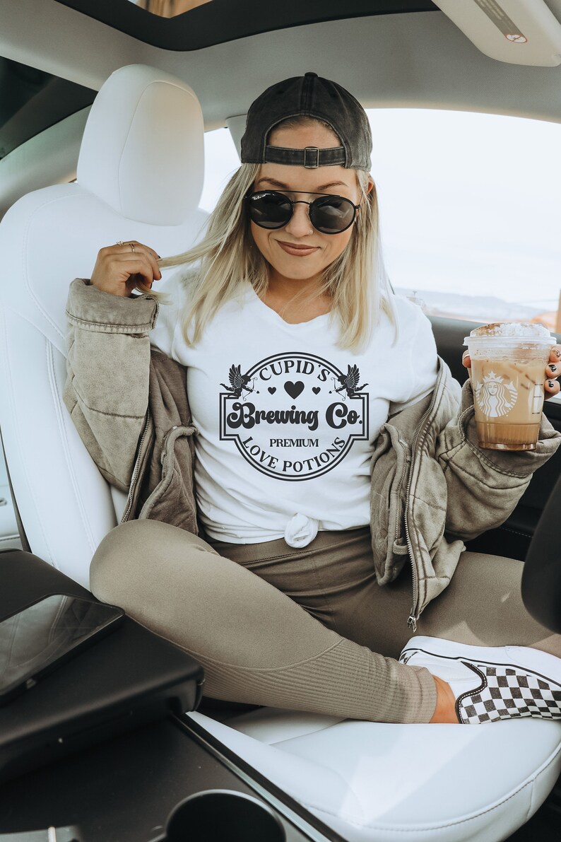 May include: White t-shirt with a black graphic that says "Cupid's Brewing Co. Premium Love Potions". The graphic features a heart and two winged cupids.