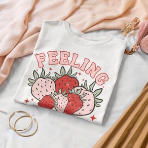 Puede incluir: Camiseta blanca con el texto "Feeling" en letras rosas y un gr&aacute;fico de fresas rojas y rosas.