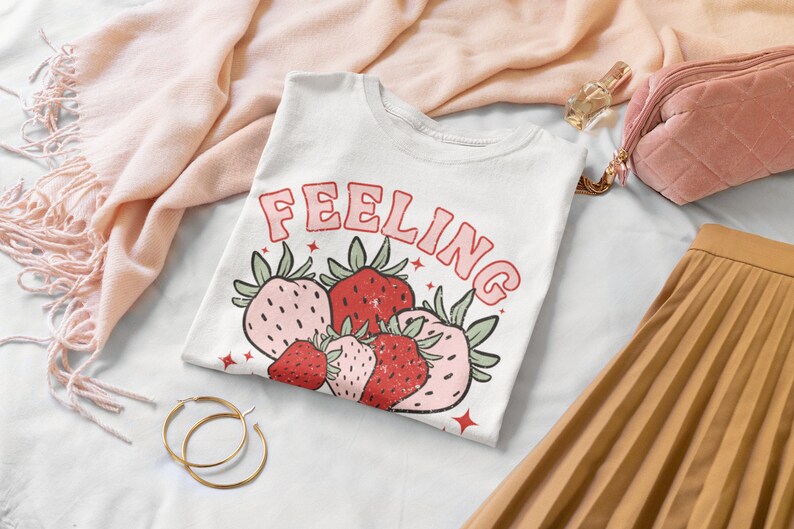 Puede incluir: Camiseta blanca con el texto "Feeling" en letras rosas y un gr&aacute;fico de fresas rojas y rosas.