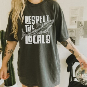 a woman wearing an oversize shirt shirt.  the shirt features a graphic of a shark and the words "respect the locals" in a retro text.  she is standing in front of a styled background