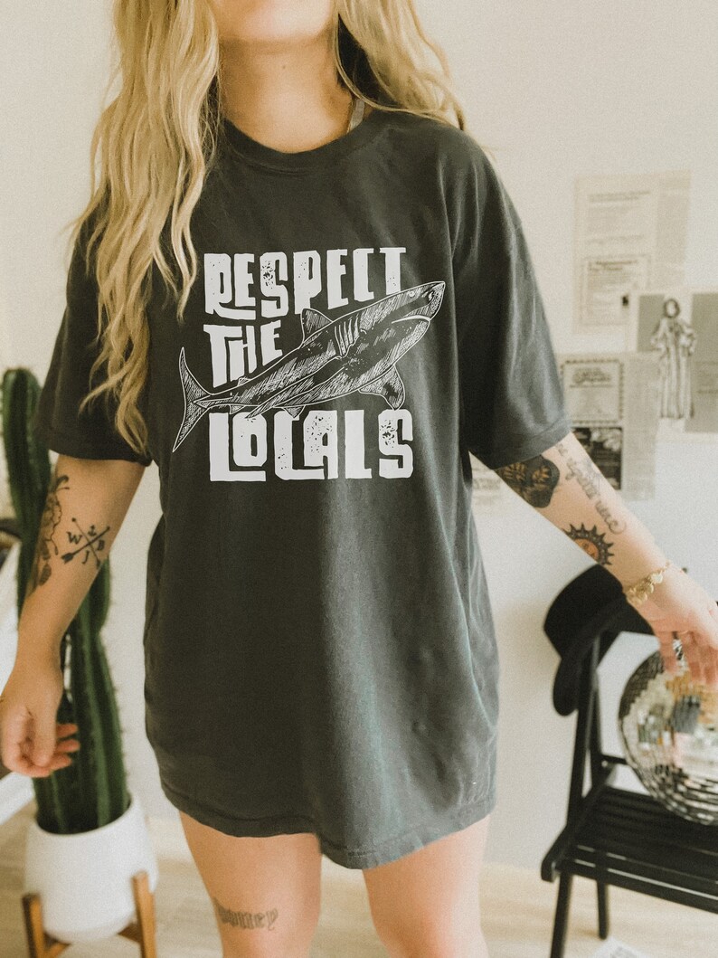 a woman wearing an oversize shirt shirt.  the shirt features a graphic of a shark and the words "respect the locals" in a retro text.  she is standing in front of a styled background